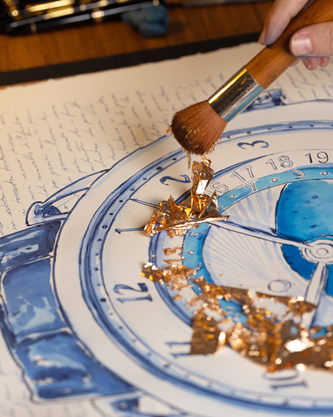 A broad brush holding gold leaf above the De Bethune dial drawing.