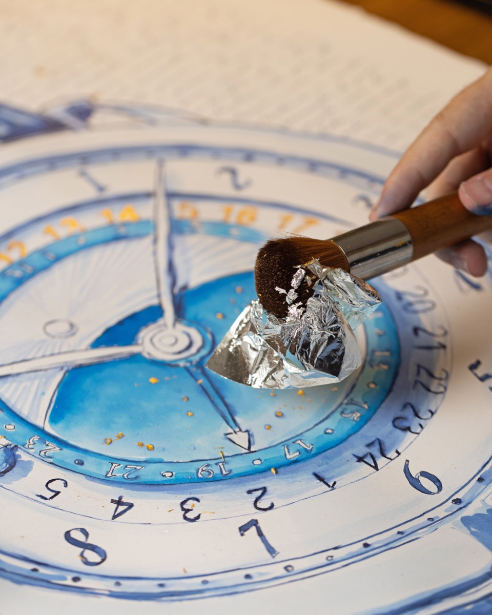 A broad brush holding reflective leaf above the blue De Bethune dial drawing.