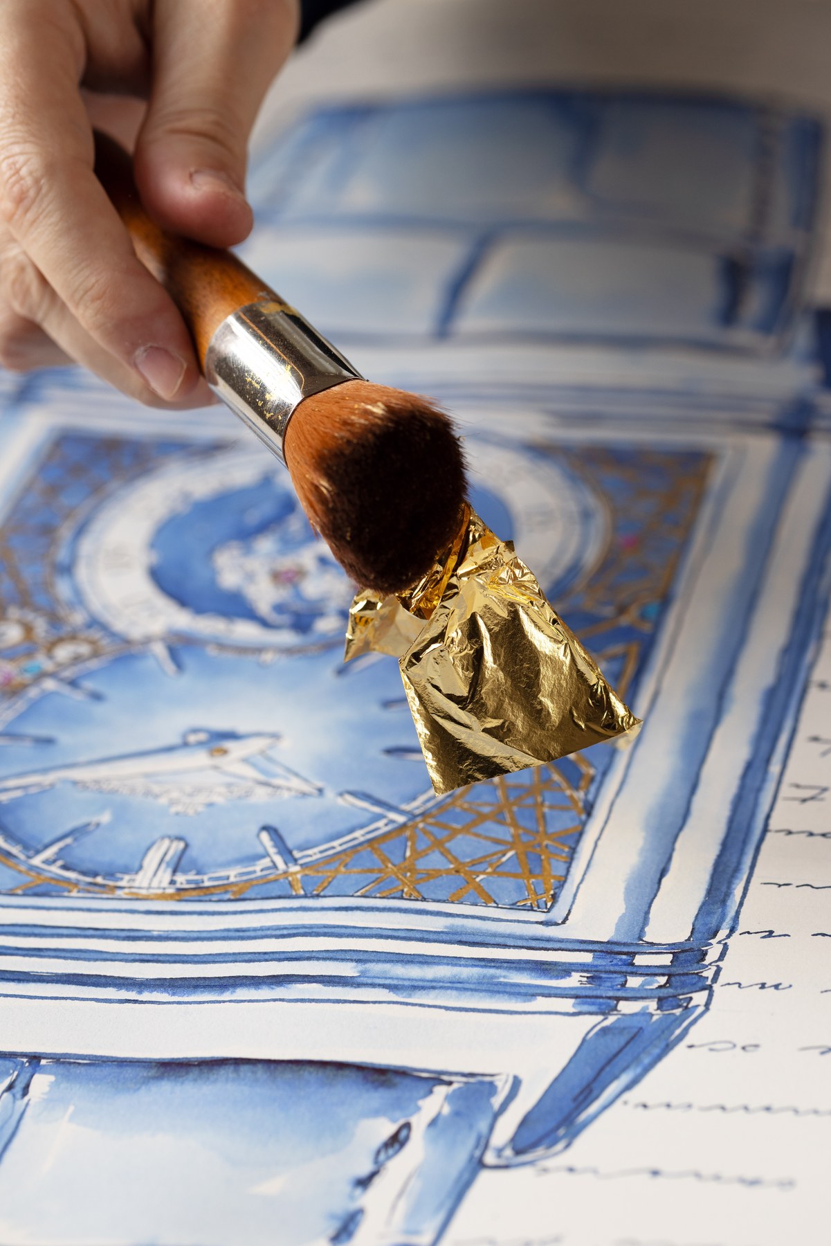 A broad brush holding gold leaf above the blue-ink watch drawing.