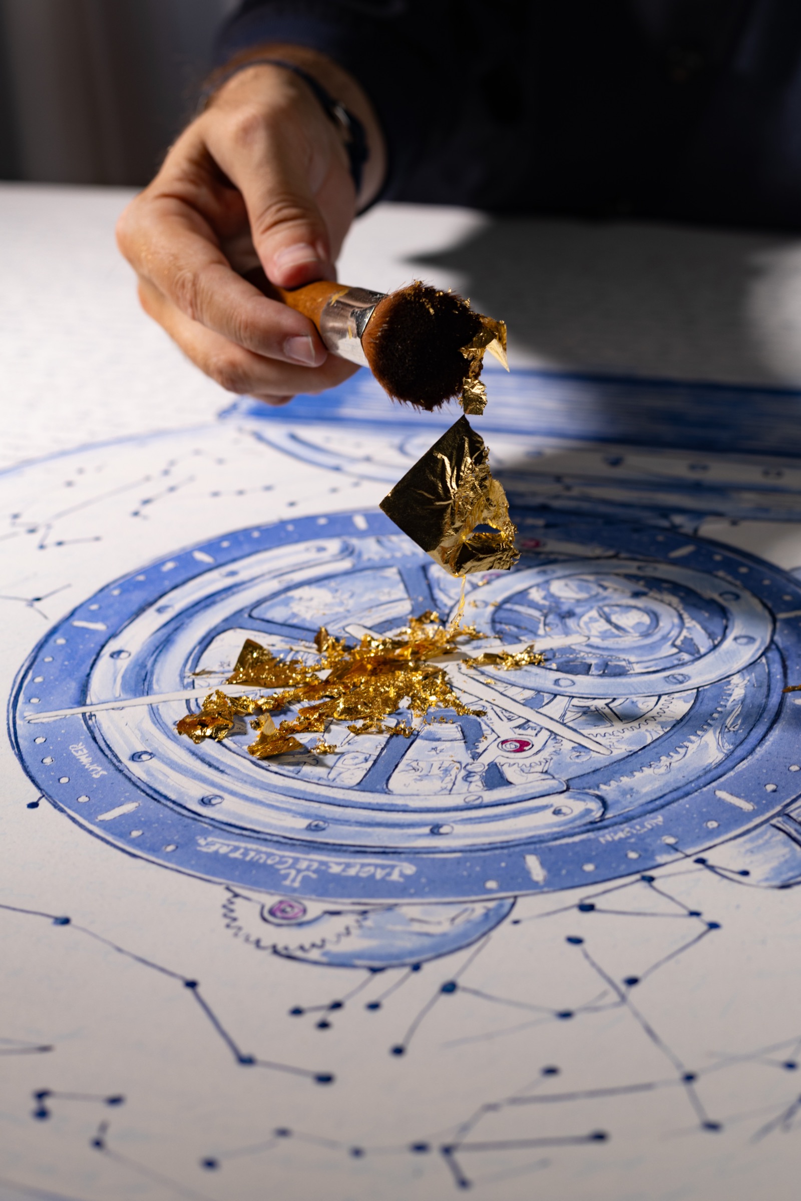 A brush lifting gold leaf above the centre of the Atmos drawing.